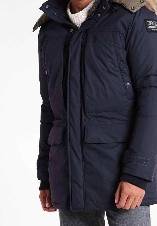 Mörkblå vadderad jacka med avtagbar huva, utrustad med två fickor fram, tryckknappar och en strukturerad stickad krage.