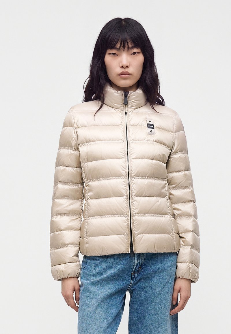 Jeune femme aux cheveux noirs portant une veste matelassée beige clair et un jean bleu, debout devant un fond uni.