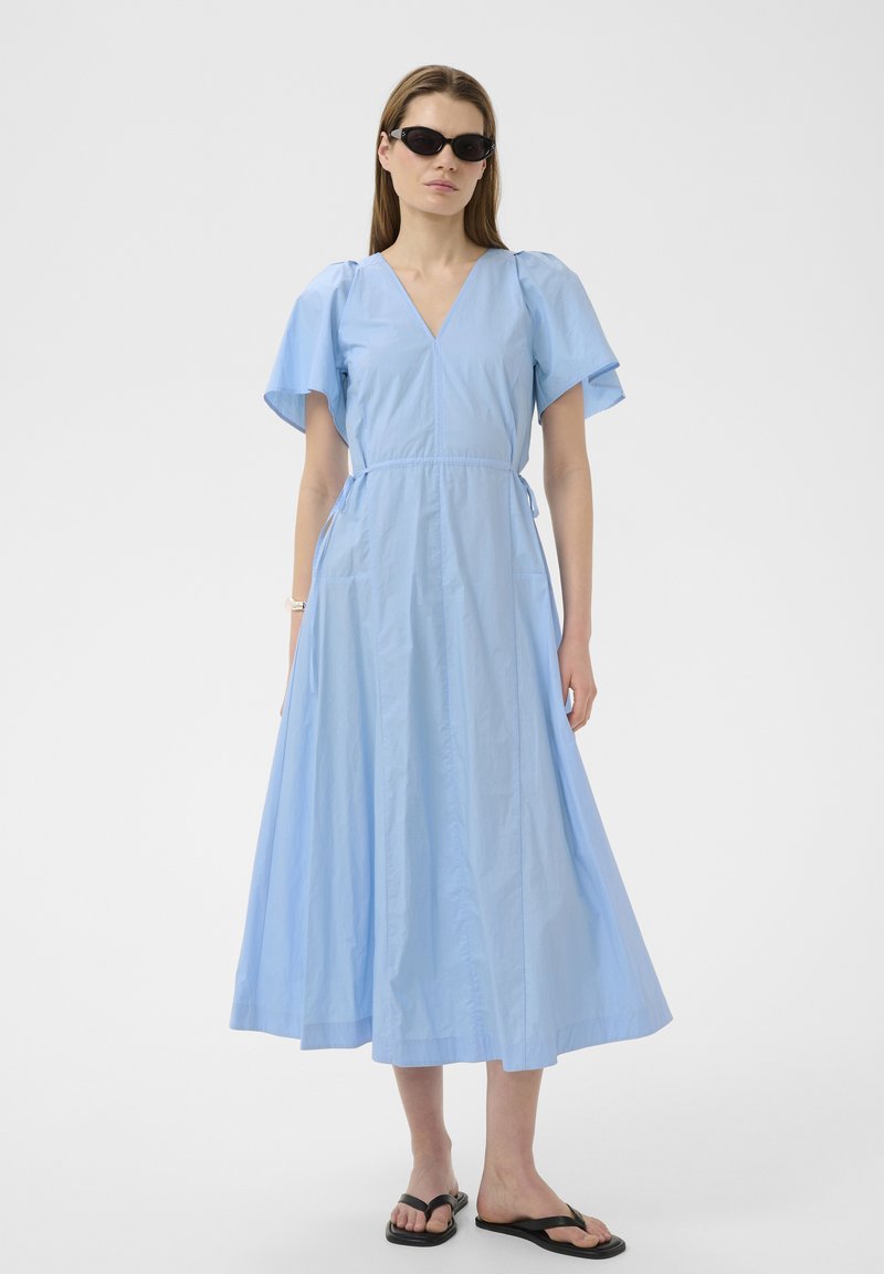 Femme portant une robe midi bleu clair à col en V avec manches flottantes, lunettes de soleil noires, tongs noires, debout devant un fond blanc uni.