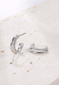 Pendientes de aro plateados con detalles de cuentas, que presentan un acabado suave y una forma curvada, colocados sobre una superficie texturizada de color claro.