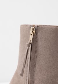 Taupe zamszowe botki za kostkę z złotym zamkiem błyskawicznym i dekoracyjną pętelką, z wyraźnym szwem wzdłuż szwów i gładką teksturą.