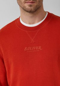 Mężczyzna w czerwonej bluzie z napisem "S.OLIVER ELEVATED SINCE 1969", nałożonej na białą koszulkę z okrągłym dekoltem.