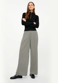 Top lungo a maniche nere a costine con zip al collo, abbinato a pantaloni a gamba larga e vita alta in un motivo houndstooth bianco e nero.