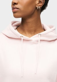 Κοντινό πλάνο ενός ατόμου που φοράει ανοιχτό ροζ φούτερ με κορδόνια, εμφανίζοντας πολλά ασημένια σκουλαρίκια και ένα κρίκο στη μύτη σ ε καφέ χρώμα δέρματος.