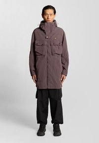 Lila, lätt, lång parka med huva, två bröstfickor och en logotyp, kombinerad med svarta lösa byxor och mörka sneakers.