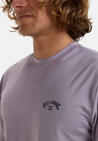 Camisa de manga corta morada hecha de una tela suave, con un logo negro en el lado izquierdo del pecho y un cuello redondo con doble costura.