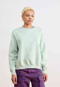 Sudadera verde menta hecha de tela suave, con cuello redondo y puños acanalados, y un pequeño logo blanco en el pecho. Combinada con pantalones cargo morados.