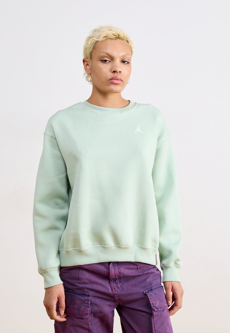 Sudadera verde menta hecha de tela suave, con cuello redondo y puños acanalados, y un pequeño logo blanco en el pecho. Combinada con pantalones cargo morados.