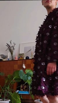 Άτομο με σκούρο μωβ φόρεμα με λουλουδάτες απλικέ, που στέκεται κοντά σε μια ξύλινη ντουλάπα διακοσμημένη με φυτά, κεριά και καδραρισμένα έργα τέχνης.