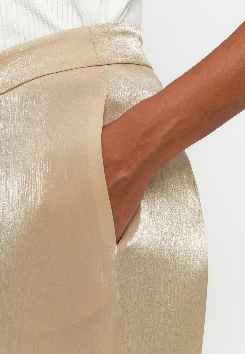 Die Hand ruht in der Tasche einer glänzenden, hellbeigen, hoch geschnittenen Hose mit glatter Textur und gesticktem Bund, getragen mit einem weißen gerippten Oberteil.
