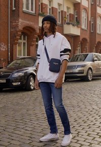 Felpa bianca con strisce nere, jeans blu e sneakers bianche. Indossa un berretto nero e ha una piccola borsa a tracolla nera. Strada acciottolata.