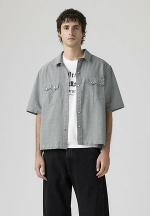 Joven con camisa de manga corta a cuadros abotonada sobre una camiseta gráfica blanca y pantalones negros, posando frente a un fondo liso.