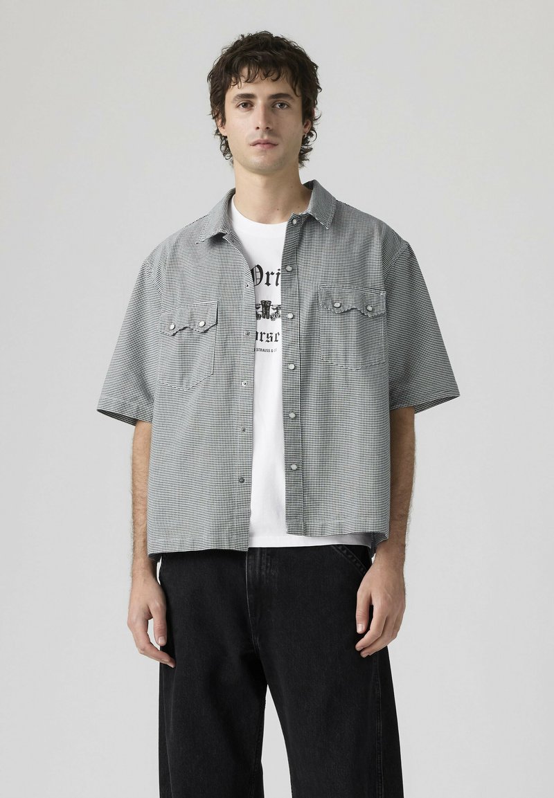 Giovane uomo con indosso una camicia a quadri a maniche corte sopra una t-shirt bianca con stampa grafica e pantaloni neri, in piedi davanti a uno sfondo semplice.