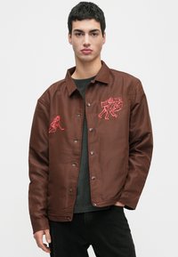 Jovem a usar uma jaqueta castanha com botões, decorada com figuras bordadas em vermelho, combinada com uma camisa escura e calças pretas, contra um fundo simples.