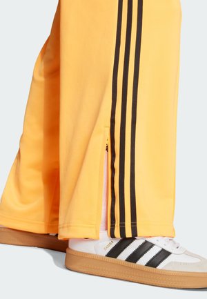 Sárga sportnadrág fekete oldalsávokkal és cipzárokkal az alján, fehér Adidas Samba cipővel fekete csíkokkal párosítva.