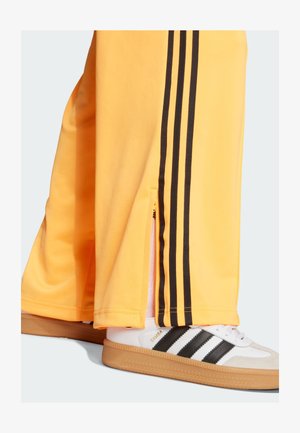 Kollaste spordipükste külgede mustade triipudega ja soontega lõpus, millele on sobitatud valged Adidas Samba kingad mustade triipudega.