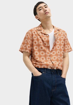 Mężczyzna ubrany w pomarańczową koszulę z krótkim rękawem z białymi wzorami słońca na białym t-shircie, w połączeniu z ciemnoniebieskimi jeansami o szerokich nogawkach, ręce w kieszeniach.