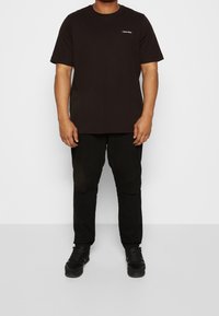 Svart t-shirt med kort ärm och rund halsringning, med en logotyp på bröstet, tillsammans med svarta slitna jeans och svarta sneakers.