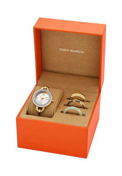 Montre Tory Burch ton or avec cadran blanc et trois lunettes colorées interchangeables dans un coffret ouvert orange et beige.