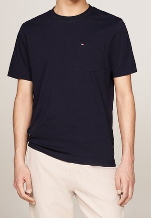 Mężczyzna ubrany w granatowy T-shirt z krótkim rękawem i małą kieszenią na piersi oraz beżowe spodnie na neutralnym tle.