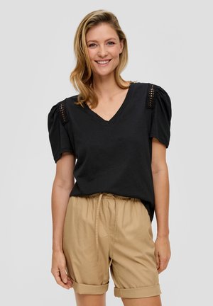 Schwarzes V-Ausschnitt-T-Shirt mit Puffärmeln, das strukturierte Akzente an den Schultern aufweist, kombiniert mit hellbeigen Shorts mit Kordelzug.