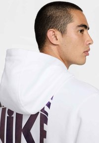 Sudadera blanca con un logo de Nike en morado en la parte posterior, con capucha ajustable y una tela suave y texturizada. Costuras detalladas visibles.