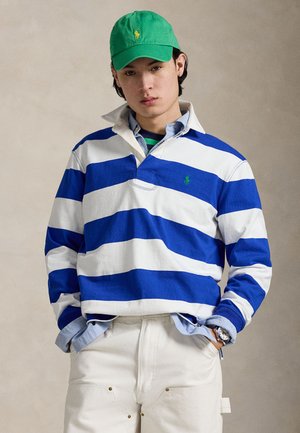 Maglia da rugby a righe bianche e blu con logo ricamato verde, indossata sopra una camicia azzurro chiaro, abbinata a pantaloni color crema.