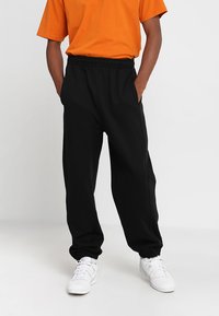 Pantalones deportivos negros con cintura y puños elásticos, que cuentan con bolsillos laterales. Llevados con una camiseta naranja y zapatillas blancas.