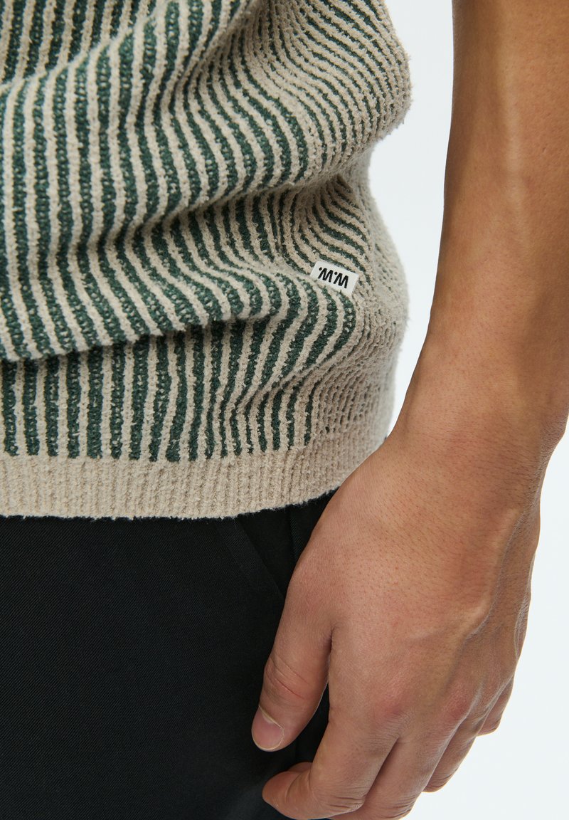 Knitted sweater featuring alternating green and beige stripes, textured fabric, with a small tag visible on the left side. Black pants partially shown.