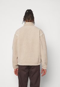 Beige fleecetröja med hög krage, avslappnad passform och texturerad yta; har sidfickor och ribbad fåll; bärs med mörka byxor.