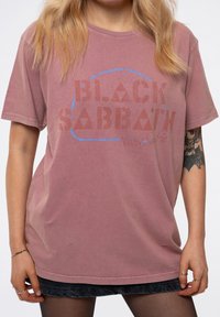 Oversized, short-sleeve pink t-shirt featuring "BLACK SABBATH" in distressed red and blue text. Crafted from soft cotton fabric.