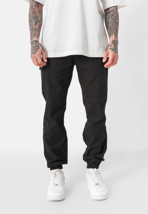Schwarze Cargo-Hosen mit schmalem Schnitt, elastischen Bündchen, Seitentaschen und einem strapazierfähigen Stoff; kombiniert mit weißen Sneakern und einem lockeren weißen Hemd.