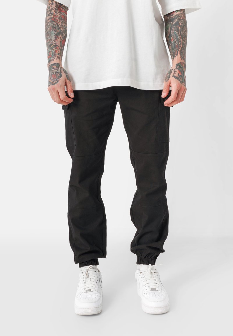 Zwarte cargobroek met een tapered fit, elastische boorden, zijzakken en een duurzame stof; gecombineerd met witte sneakers en een losse witte shirt.