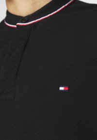 Polo noir en tissu texturé avec un col arrondi, rehaussé de surpiqûres rouges et blanches, et un petit logo brodé.