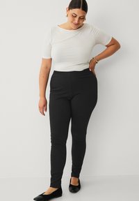Sorte stræk-leggings med en glat tekstur og høj talje, kombineret med en tætsiddende hvid top og sorte flade sko. Simplet design.