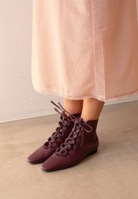 Botas de piel burdeos hasta el tobillo con punta afilada y cordones decorativos. Llevadas con una falda rosa claro con ribete de encaje. Textura suave.