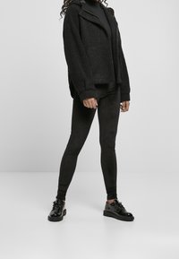 Veste en polaire noire avec fermeture éclair et capuche, associée à des leggings noirs et des chaussures noires brillantes, sur fond neutre.