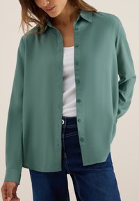 Chemisier vert à boutons en tissu léger, avec un col classique et des manches longues, associé à un jean en denim bleu et un t-shirt blanc.