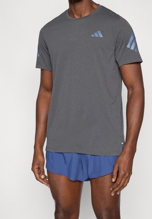 Homme portant un t-shirt gris à manches courtes avec des logos bleus et un short de sport bleu, debout devant un fond uni.