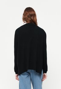 Čierny pletený cardigan s voľným strihom, dlhými rukávmi, asymetrickým lemom a rebrovanými akcentmi. Tkanina vyzerá mäkko a má textúru.