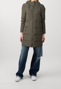 Kaki puffjacka med huva, dragkedjestängning och två sidofickor. Har horisontell sömnad och en lätt, något glänsande yta.