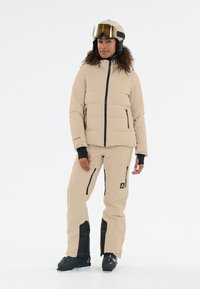 Beige isolerad skidjacka med svart dragkedja, matchad med beige byxor som har svarta detaljer och fickor med dragkedja. Svarta skidstövlar.