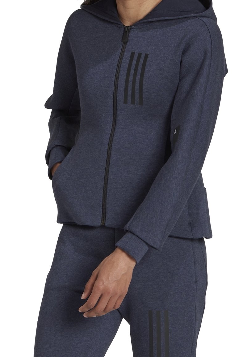 Personne portant un sweat à capuche zippé bleu foncé et un pantalon assorti avec trois bandes noires verticales sur la poitrine et la cuisse, mains dans les poches.