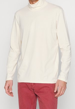 Krémový rolák s dlouhým rukávem vyrobený z měkké tkaniny, s uvolněným střihem a jednoduchým designem. V náhledu spárován s červenými kalhotami.