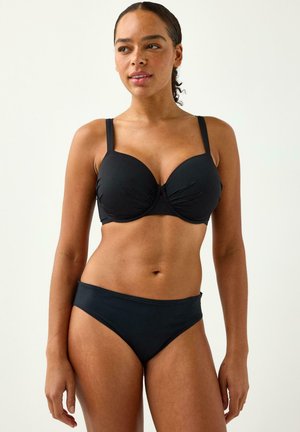 Donna in piedi che indossa un reggiseno nero con ferretto e slip bikini neri abbinati, su uno sfondo chiaro uniforme.