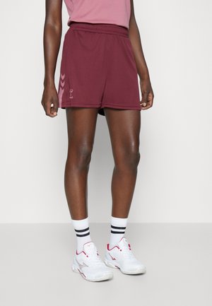 Person wearing maroon athletic shorts, pink top, white socks with black stripes, and white sneakers with pink accents standing on gray floor.