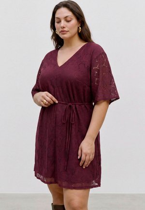 Mujer con un vestido de encaje burdeos de manga corta con escote en V y cinturón en la cintura, de pie frente a un fondo liso y claro.