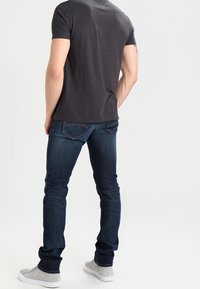 Dunkelblaue Jeans im Slim-Fit mit Gesäßtaschen und einer dezenten Waschung. Getragen mit einem dunkelgrauen T-Shirt und grauen Sneakers.