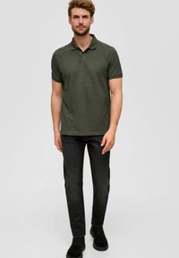 Polo vert foncé à manches courtes, col côtelé et logo. Porté avec un jean foncé et des chaussures noires à lacets. Fond neutre.