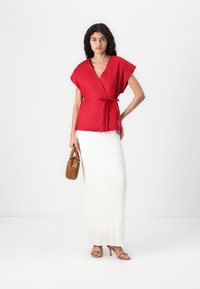 Top rosso incrociato con maniche corte, annodato in vita, abbinato a una maxi gonna bianca texturizzata e una borsa marrone. Sandali con tacco rosa completano il look.